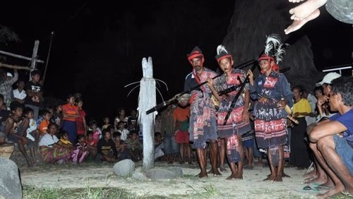 Three men in traditional West Sumba ceremonial dress and patterned textiles holding long spears during a nighttime ritual called Bijalungu Hiu Paana, with a crowd of onlookers in the background.
