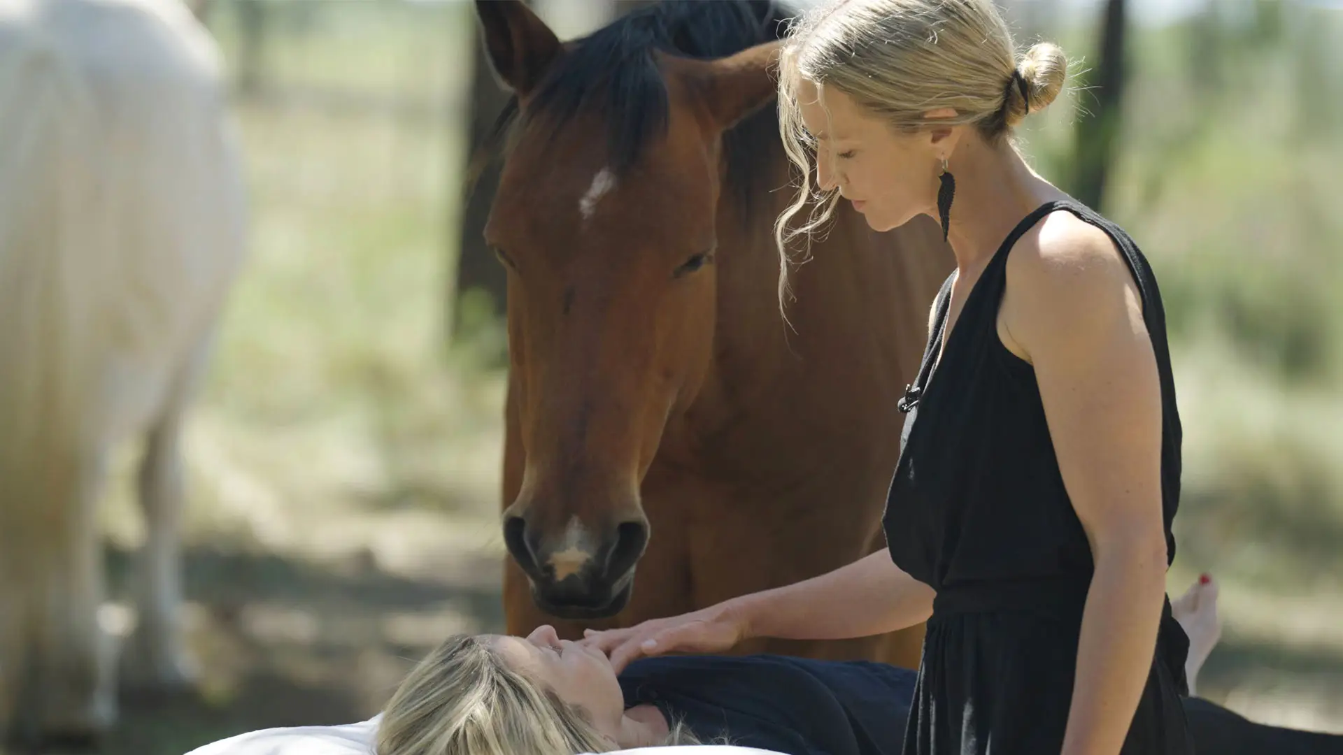 A blonde woman in a black dress performing an equine therapy session at NIHI Sumba, gently placing her hand on a person lying down while a brown horse stands calmly in the background.