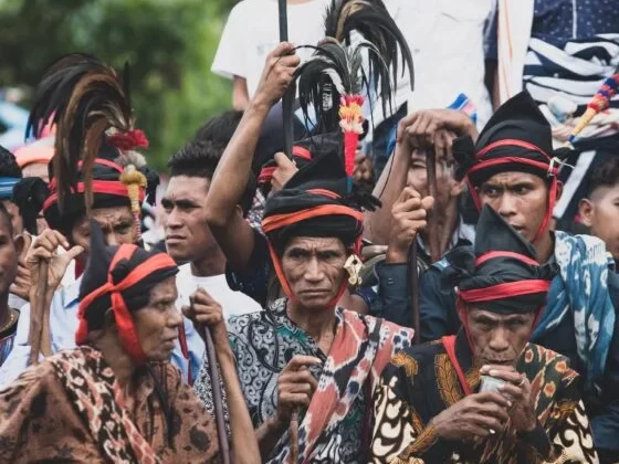 A close-up shot of a group of men in traditional Sumba attire, wearing black headcloths wrapped with red fabric and topped with tall, dark feathers. They are dressed in intricate, patterned ikat fabrics, and several men hold long wooden spears or staffs while looking forward with focused expressions.
