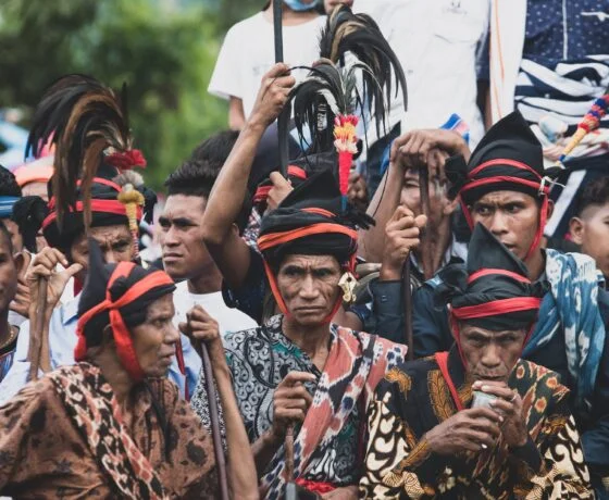 A close-up shot of a group of men in traditional Sumba attire, wearing black headcloths wrapped with red fabric and topped with tall, dark feathers. They are dressed in intricate, patterned ikat fabrics, and several men hold long wooden spears or staffs while looking forward with focused expressions.