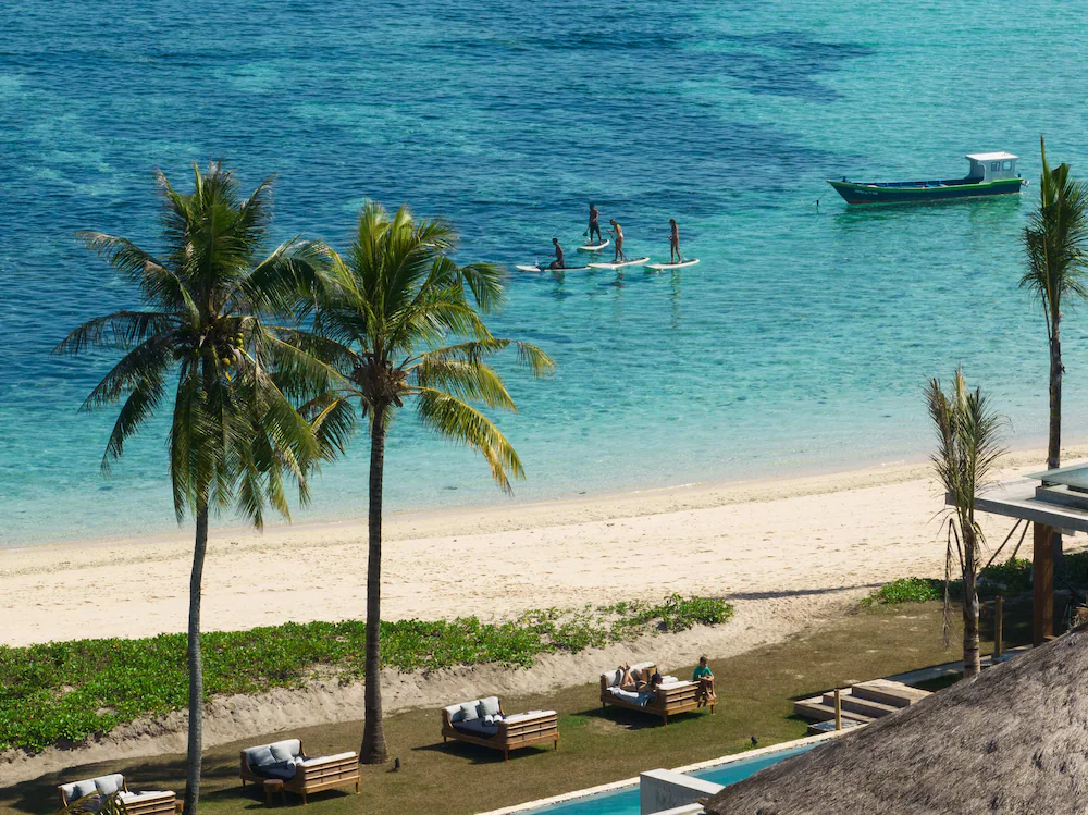 Palm trees line a sandy beach with turquoise water where people paddleboard and a boat floats in Sumba, Indonesia.