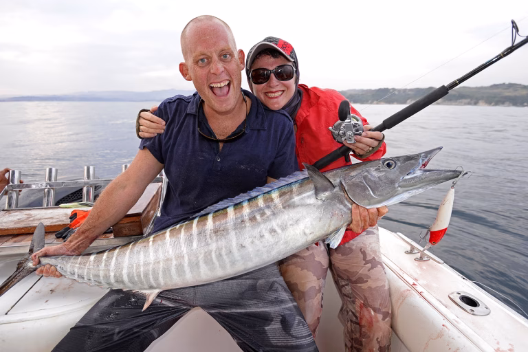 Two smiling people on a boat hold a large silver wahoo fish with white stripes, against a backdrop of ocean and distant land.