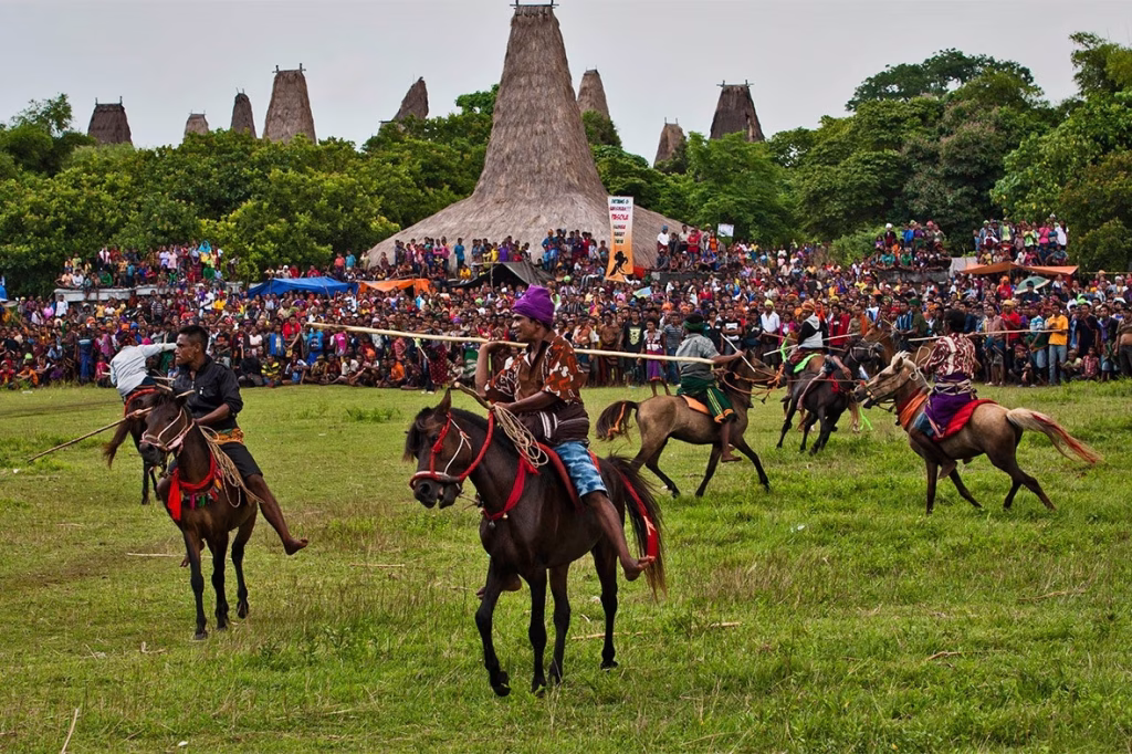 Sumba men on horseback joust with spears in a green field before a large crowd and traditional thatched-roof buildings.