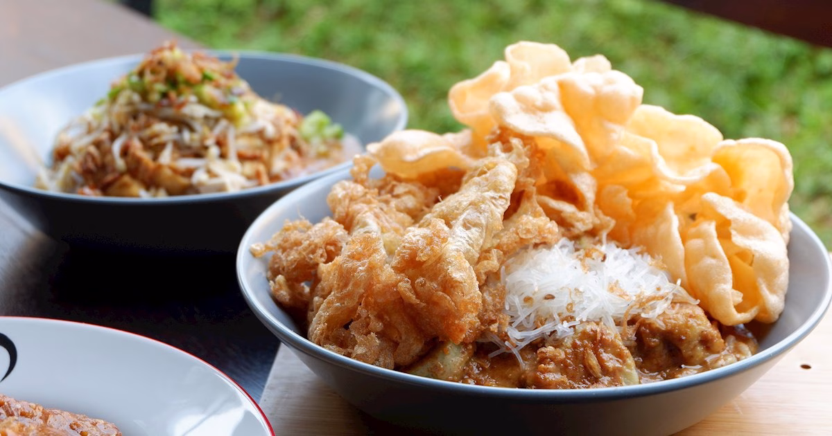 A close-up of a bowl containing a traditional Sumba food dish featuring a creamy base topped with thin rice vermicelli, large pieces of crispy fried egg or tofu, and a side of light, airy crackers. In the background, another bowl shows a similar dish garnished with fried shallots and green onions.
