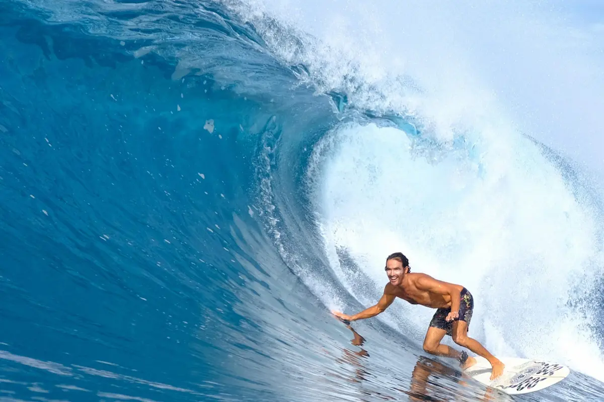 A smiling man with dark hair and board shorts is crouched on a white surfboard, riding inside a large, deep blue barreling ocean wave.