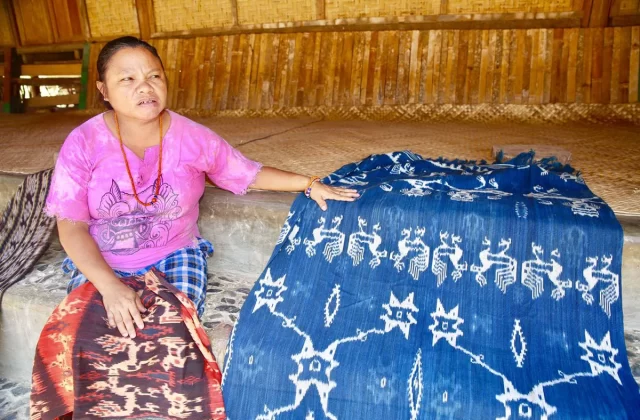 A woman in Sumba sits beside a blue ikat textile with white designs, inside a building with woven walls.