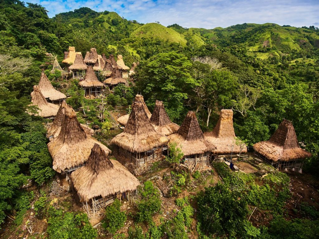 An aerial, high-angle view of a traditional Sumba village built into a lush, green hillside. The village consists of numerous houses with distinctive, high-peaked thatched roofs made of alang-alang grass. The structures are clustered together on the slope, surrounded by dense tropical forest and rolling green mountains under a bright sky, signifying Sumba culture.
