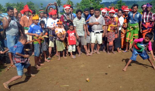A large group of people gathered outdoors in Sumba to watch a traditional top-spinning game called Maka Gasing, featuring two young participants actively launching wooden tops on a dirt ground.
