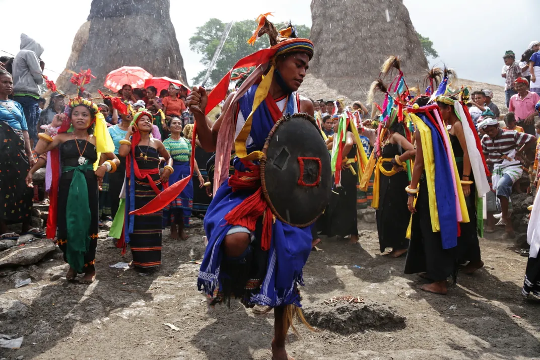During Sumba's Wulla Podu, people in colorful ceremonial attire dance with shields in a rocky outdoor setting with spectators.