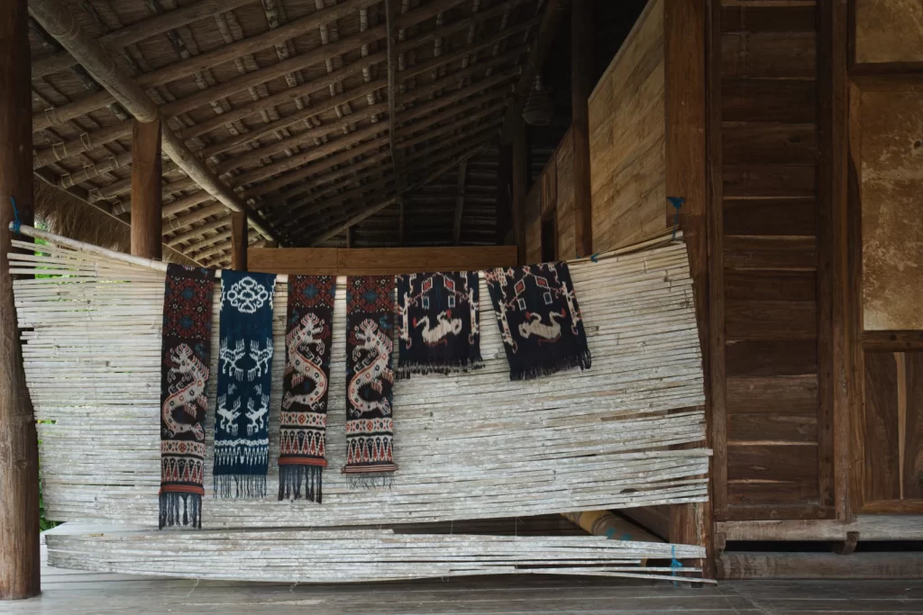 Five colorful, patterned textiles hang on a bamboo wall inside a wooden structure with a thatched roof in Sumba, Indonesia.