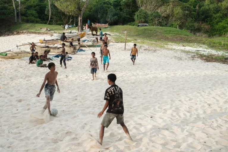 Boys are playing soccer on a white sandy beach in Sumba, Indonesia, with boats and green vegetation in the background.