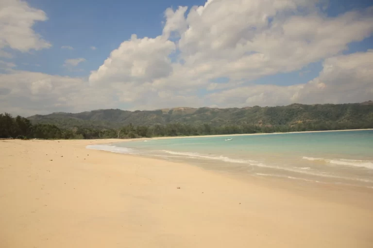 A wide sandy beach with turquoise water meets green hills under a blue sky with white clouds in Sumba, Indonesia.