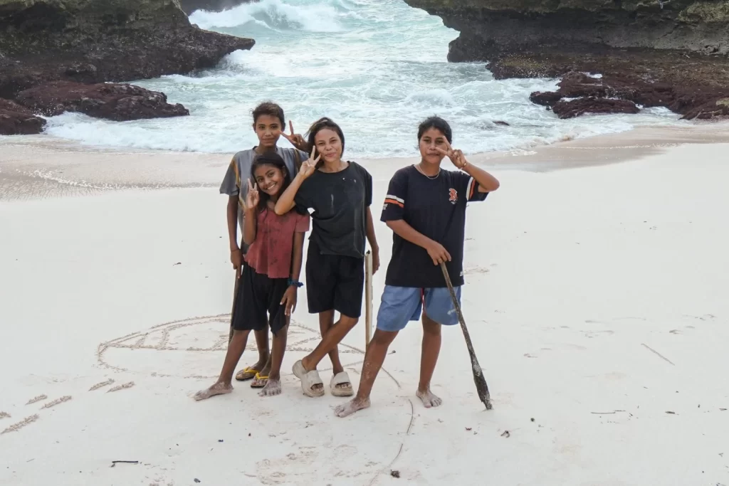 Four young people stand on a white sand beach with crashing waves and rocky cliffs in the background, making peace signs.