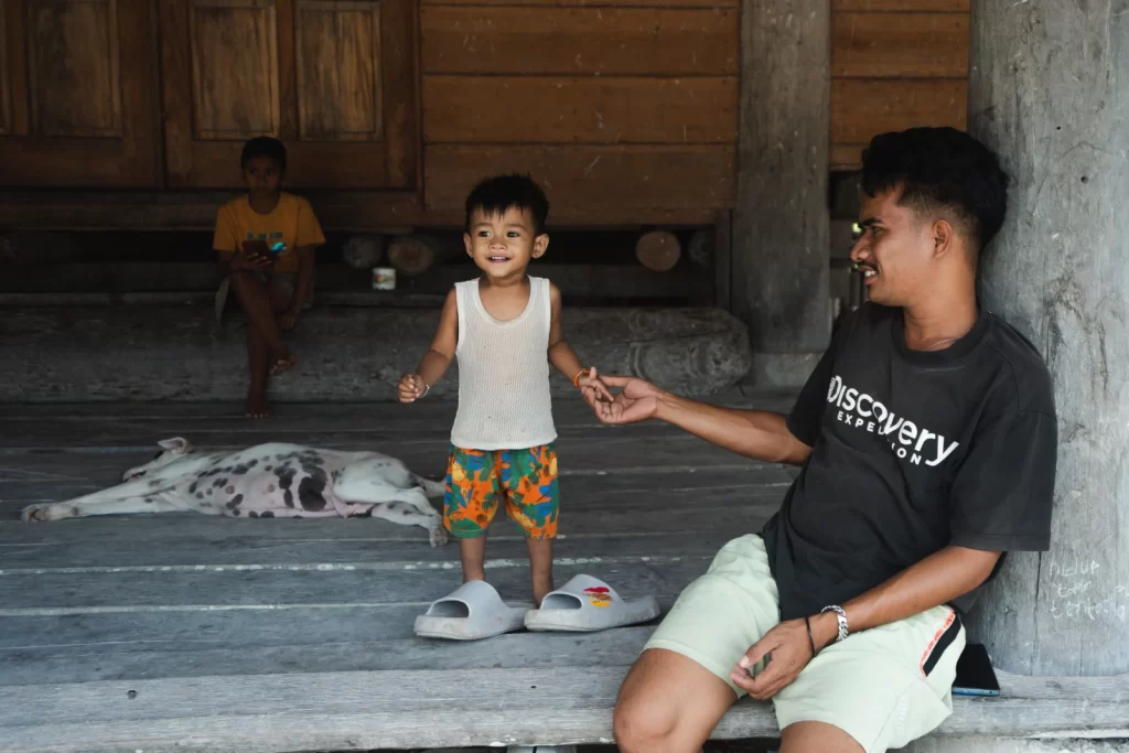 On a wooden porch, a man holds the hand of a smiling child in a white tank top and orange shorts, with another child and dog in the background.