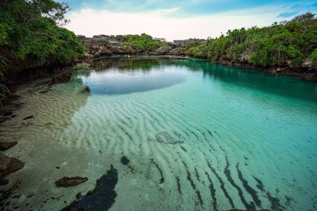 A turquoise lagoon with sandy bottom is surrounded by green vegetation and rocky cliffs under a cloudy sky in Sumba, Indonesia.