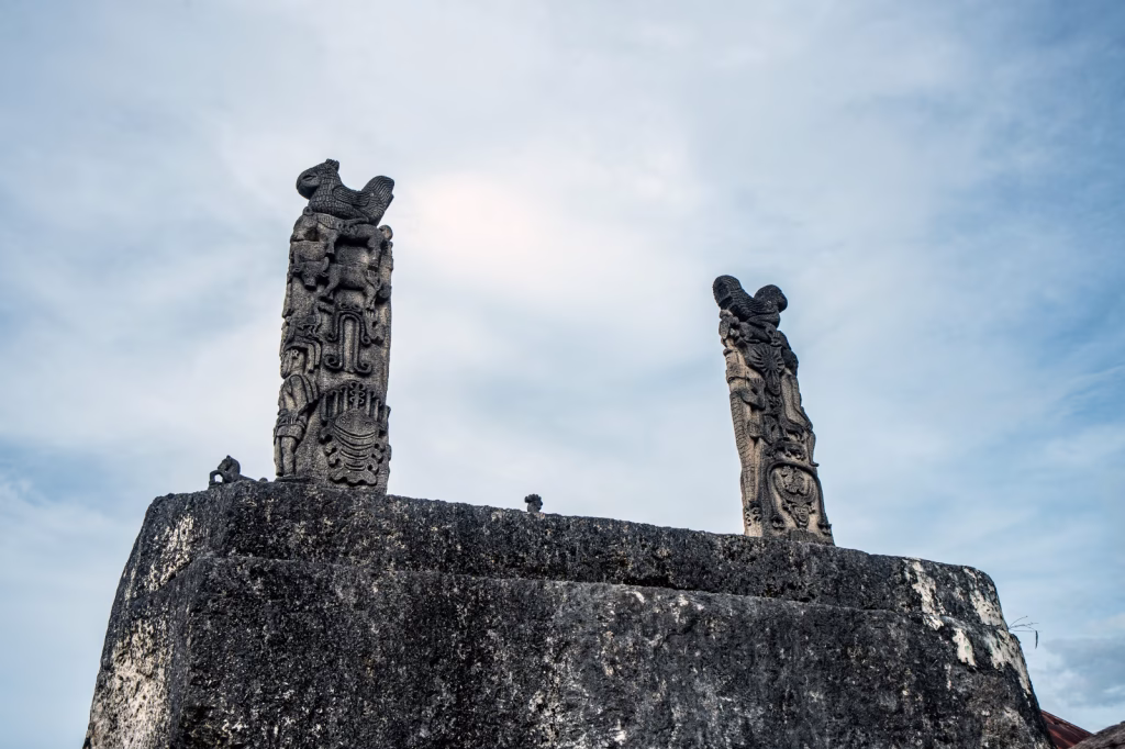 Two carved stone pillars with rooster figures stand atop a stone structure against a bright, overcast sky.