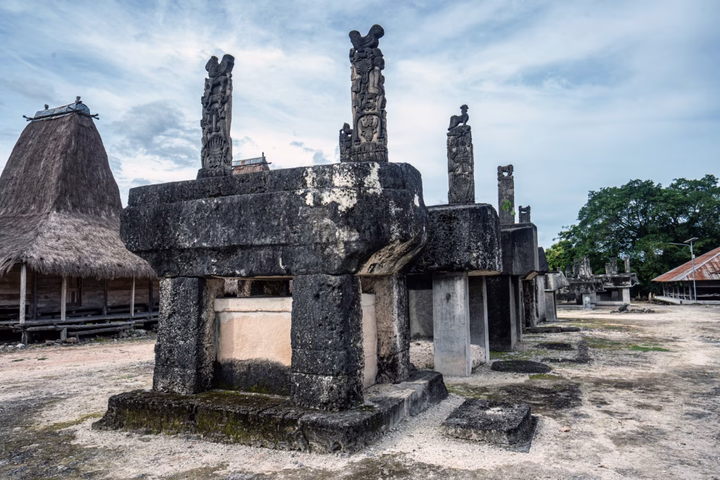 Sumba Island features stone tombs with carved animal figures, and a traditional thatched-roof house, under a bright sky. Villages in Sumba.