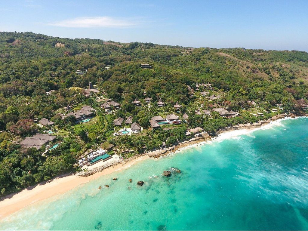 Aerial view of a resort with thatched roofs nestled between a lush green hillside and a turquoise ocean on Sumba Island.
