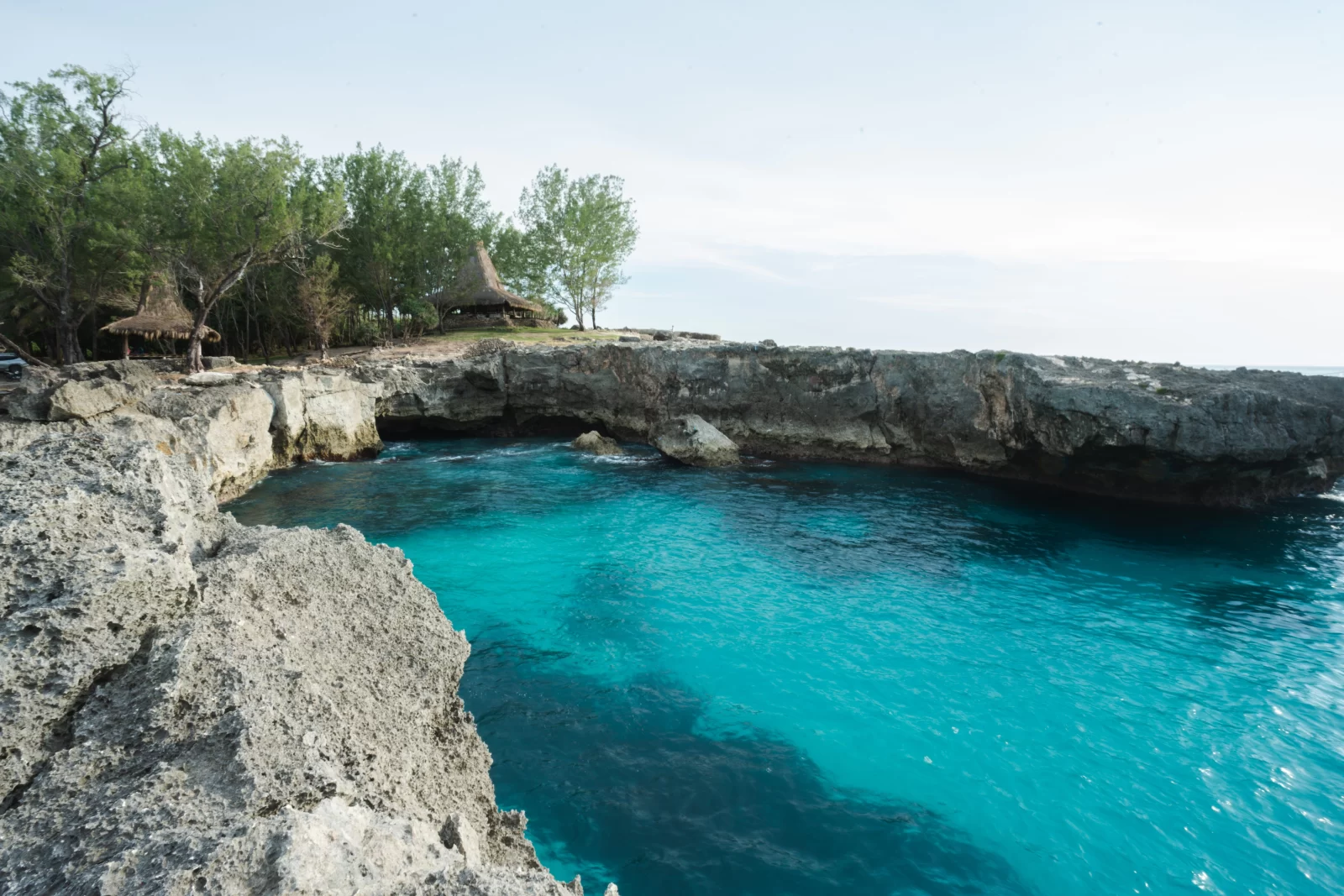 Turquoise water fills a cove surrounded by rocky cliffs and trees with thatched-roof structures on Sumba Island.
