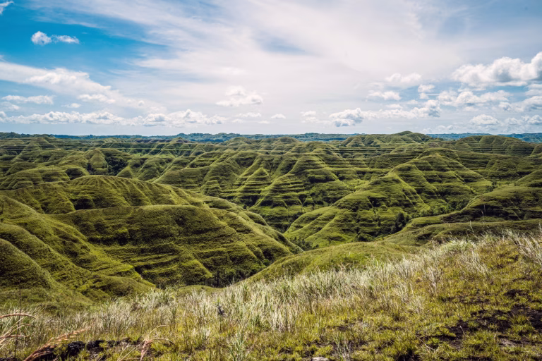Rolling green hills stretch across the landscape under a blue sky with scattered clouds in Sumba, Indonesia. Sumba geography.