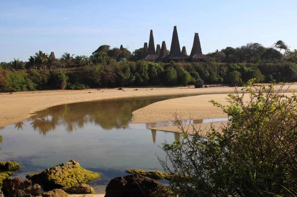 Sumba Island view shows traditional high-peaked houses beyond a sandy beach and calm water, under a blue sky. Villages in Sumba.