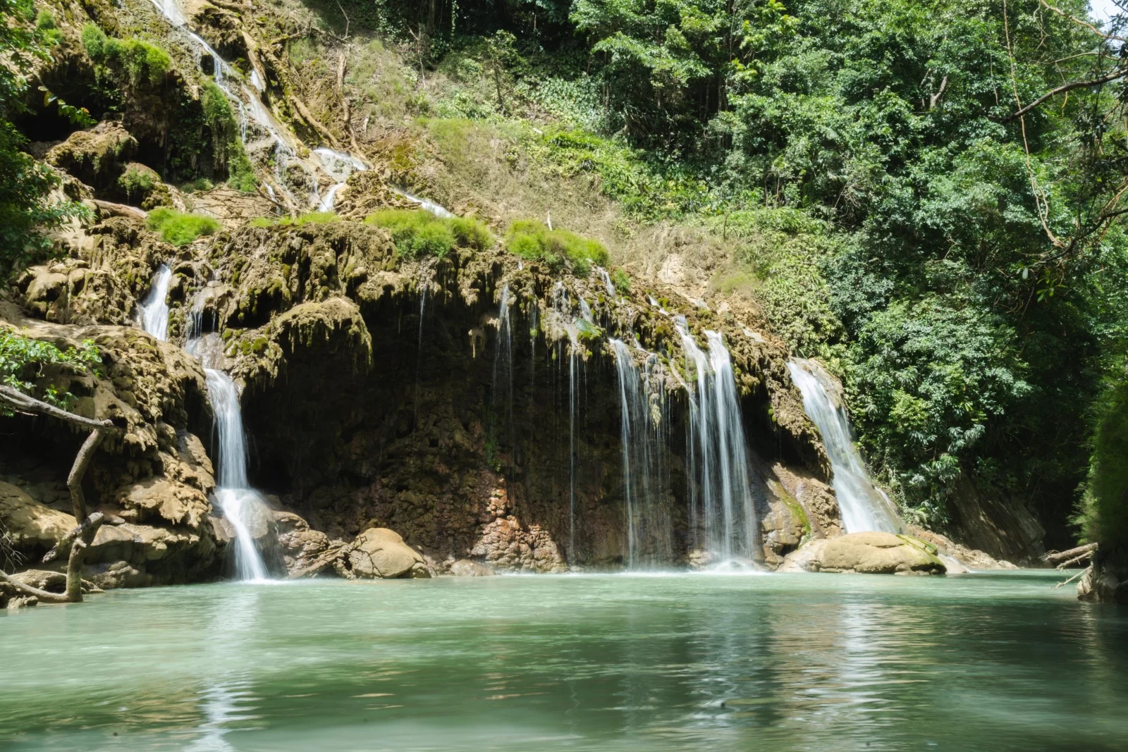 Multiple waterfalls cascade into a green pool surrounded by lush, green vegetation on Sumba Island.