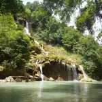 A tiered waterfall cascades into a turquoise pool, surrounded by lush green trees in a tropical setting. Day trips in Sumba.