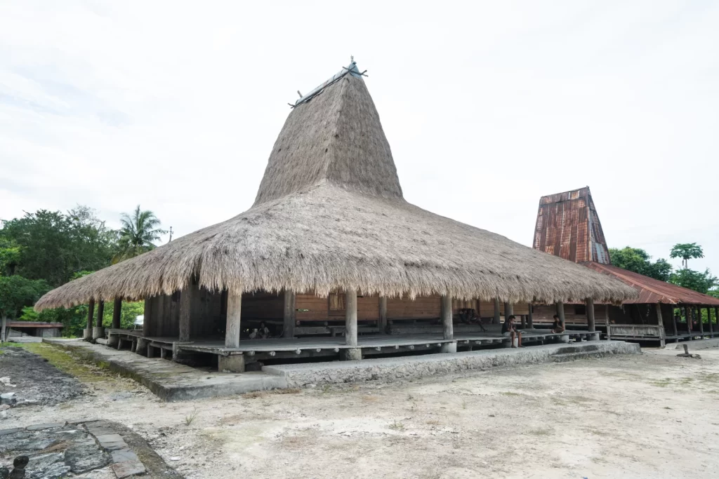A large, traditional Sumbanese house with a tall, thatched roof stands on a sandy ground under a cloudy sky. Villages in Sumba.