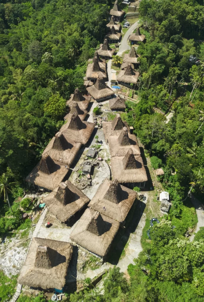 Aerial view shows traditional Sumba houses with thatched roofs in a village setting surrounded by green trees. Villages in Sumba.