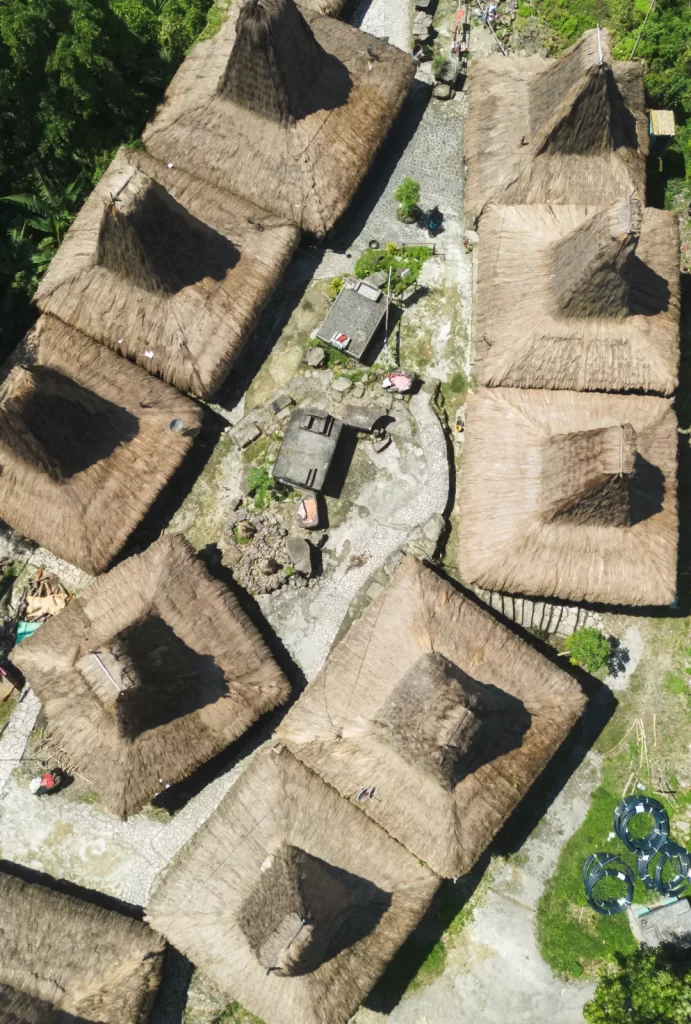 Aerial view shows traditional Sumba houses with high thatched roofs in a village setting, surrounded by green vegetation.