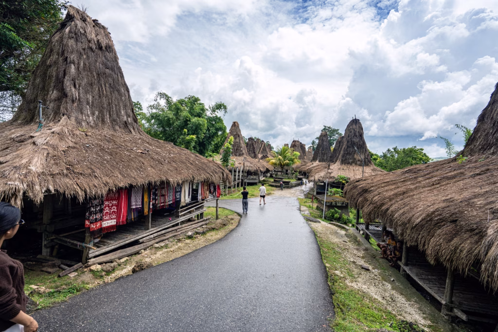 A village in Sumba, Indonesia, features traditional houses with tall, thatched roofs along a gray paved road under a cloudy sky. Villages in Sumba.