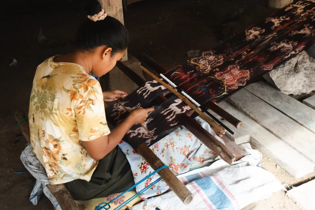A woman in a floral shirt sits weaving a dark brown and white patterned textile on a wooden loom in a rustic setting. Villages in Sumba.