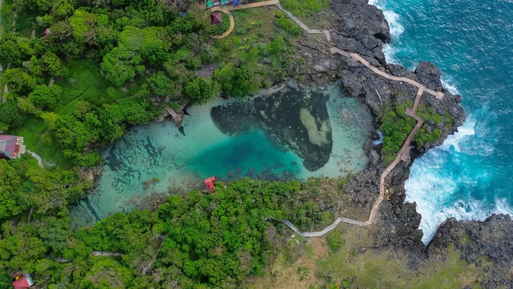 Aerial view shows a turquoise lagoon surrounded by green trees and rocky cliffs with a wooden walkway in Sumba, Indonesia.
