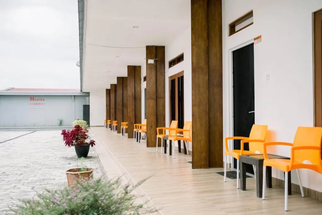 A row of orange chairs sits outside rooms with brown pillars at Morika Inn in Waingapu, Sumba, under a cloudy sky.