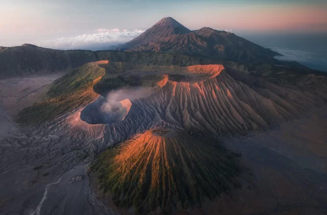 Aerial view shows three volcanoes in a mountainous landscape under a blue and pink sky at sunrise.