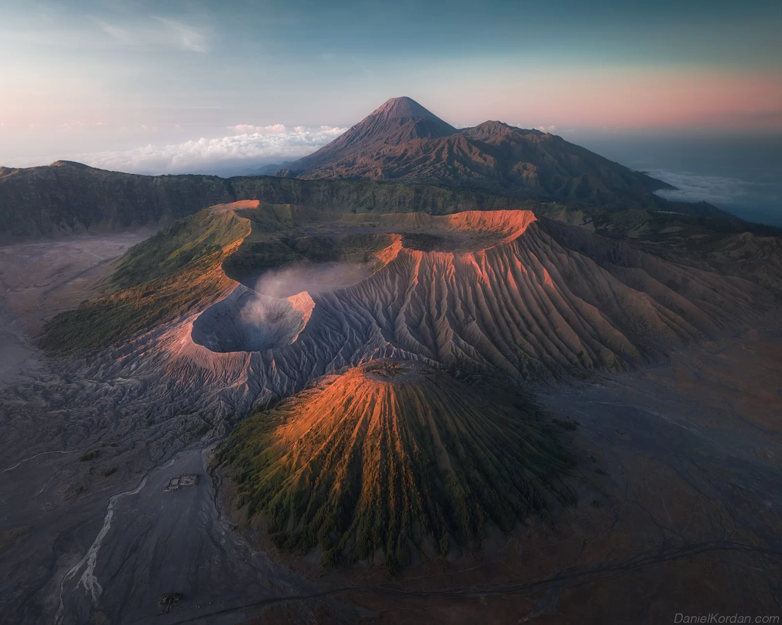 Aerial view shows three volcanoes in a mountainous landscape under a blue and pink sky at sunrise.