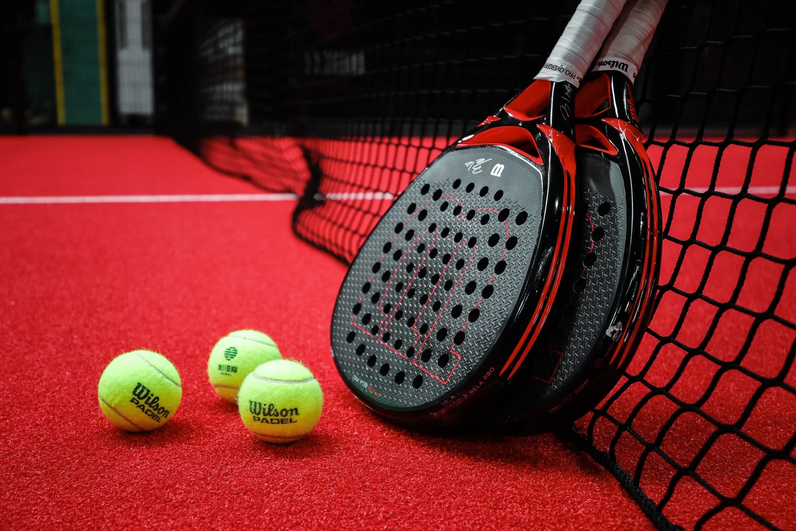 Two black and red Wilson padel rackets lean against a net with three yellow Wilson padel balls on a red surface.