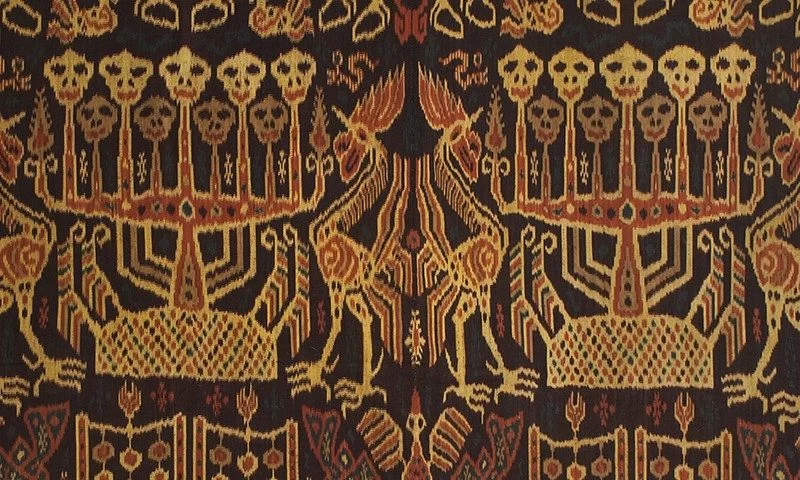 A dark brown textile features repeated patterns of stylized skulls, birds, and geometric shapes in shades of brown, orange, and gold. History of Sumba.