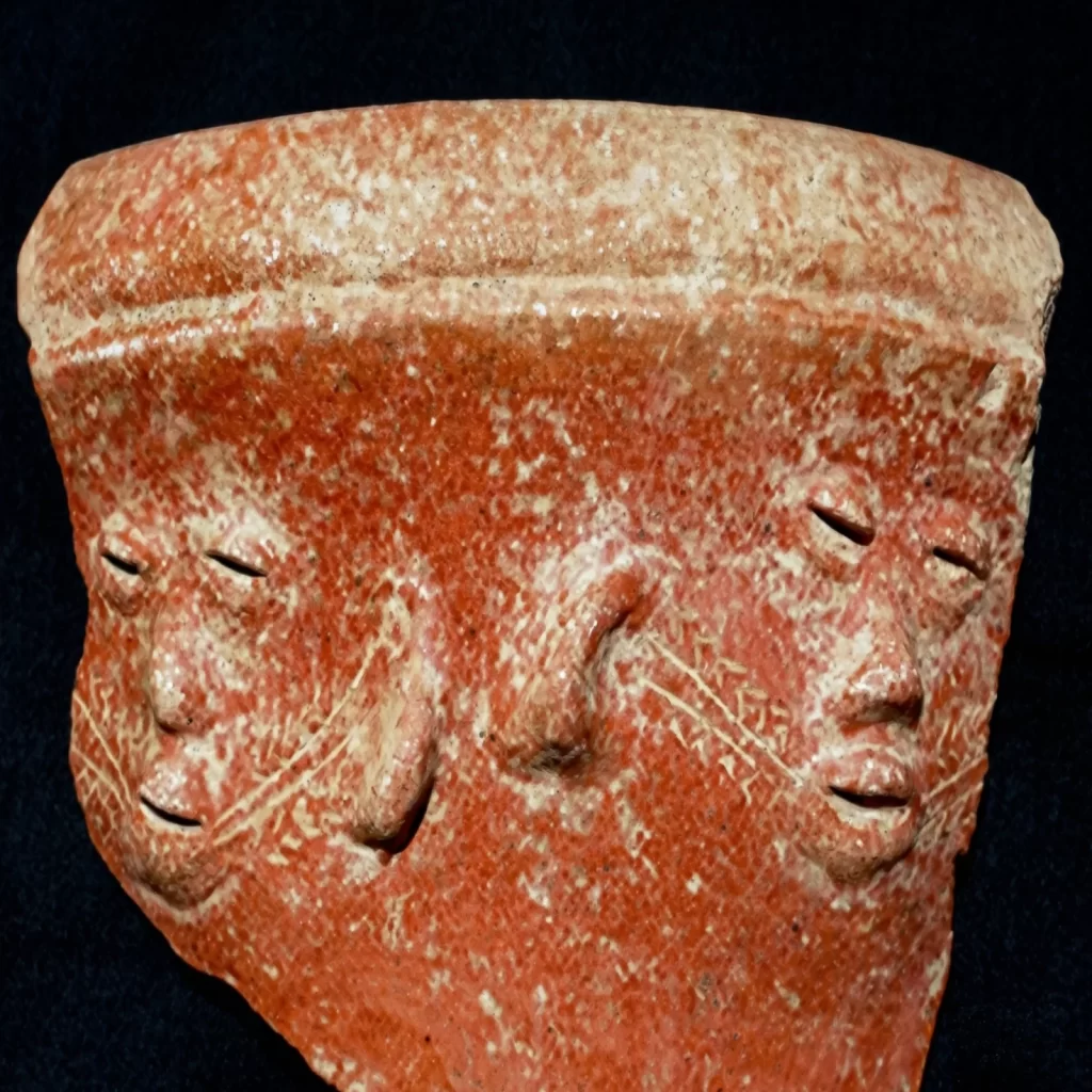 A fragment of a reddish-brown clay artifact shows two faces with closed eyes and smiling mouths, possibly from Sumba, Indonesia. History of Sumba.