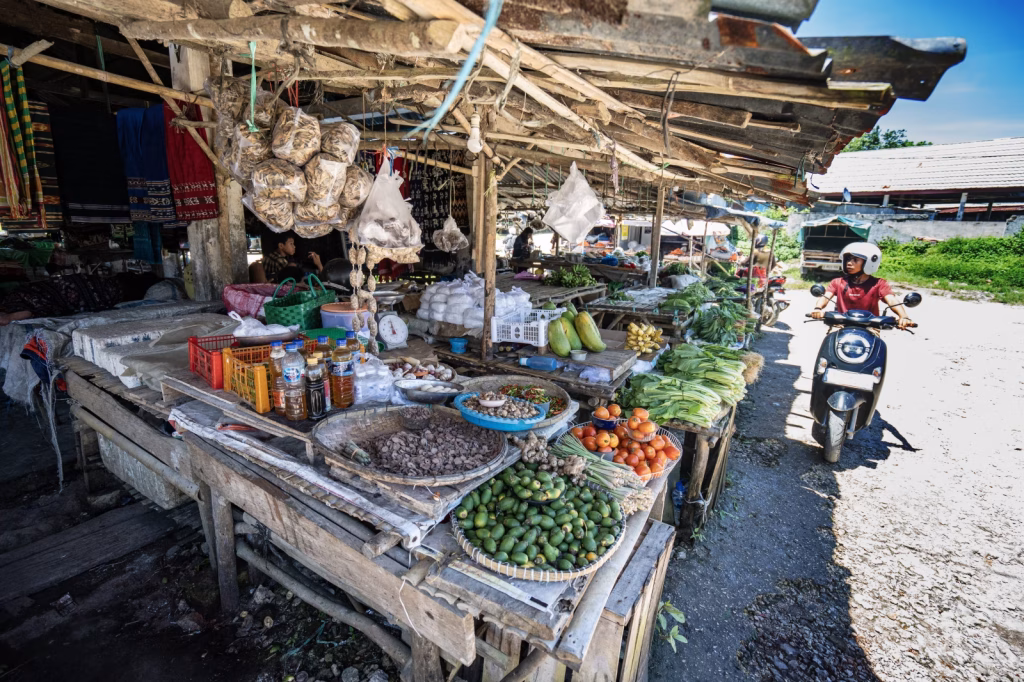 A person on a scooter rides past a market stall with produce in Sumba, Indonesia, on a sunny day.