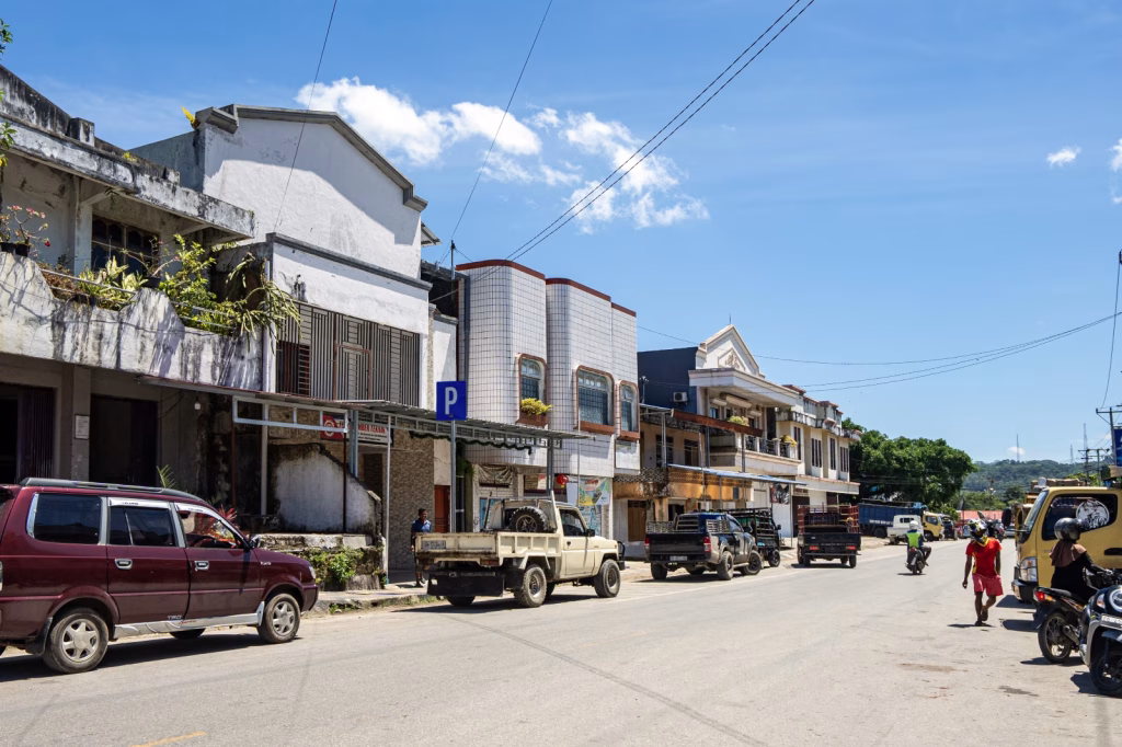 Street scene in Sumba, Indonesia, with parked cars and trucks along the roadside under a bright blue sky with scattered clouds. History of Sumba.