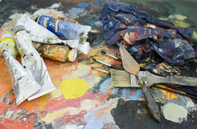 The image shows a painter's palette with tubes of paint, brushes, and a cloth, all covered in colorful paint splatters.