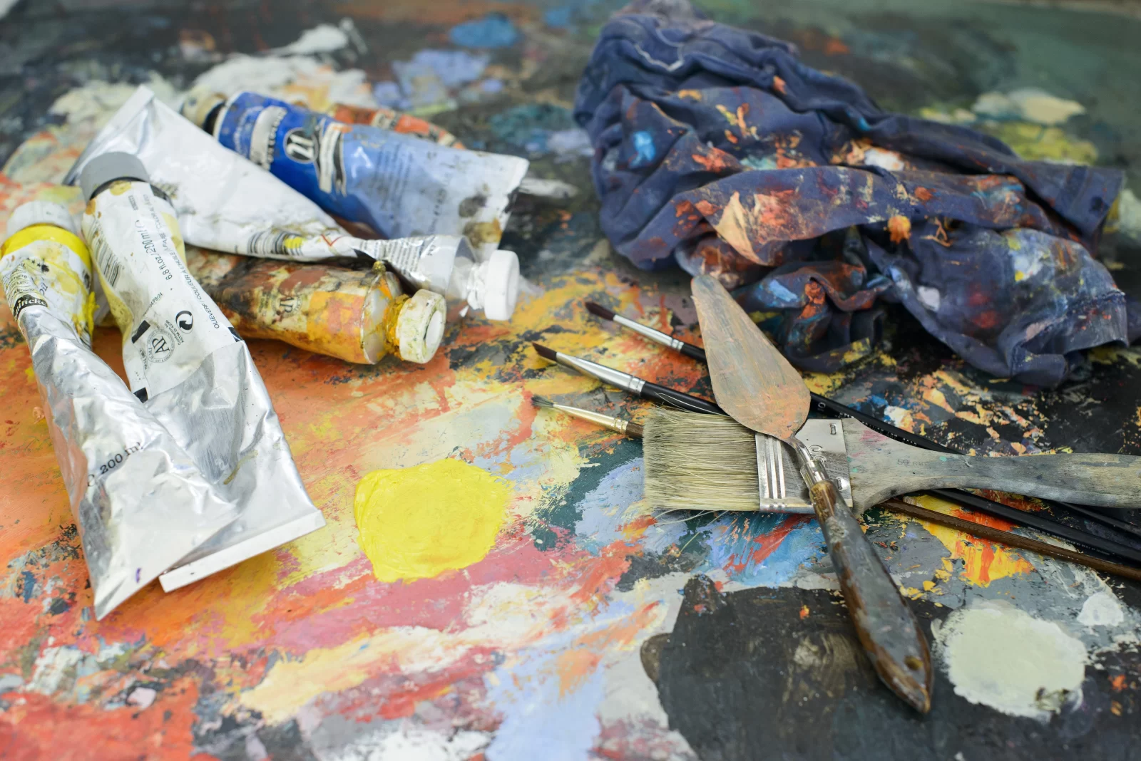 The image shows a painter's palette with tubes of paint, brushes, and a cloth, all covered in colorful paint splatters.