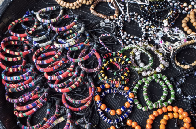 A black tray displays colorful woven and beaded bracelets in shades of red, orange, green, and brown.
