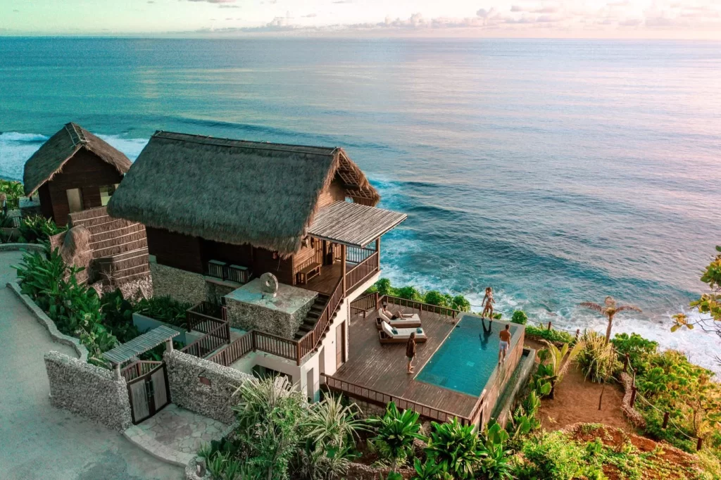Thatched-roof bungalows sit on a cliff overlooking the blue ocean, with people relaxing by an infinity pool in Sumba, Indonesia.