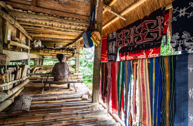 A woman sits inside a bamboo structure with colorful textiles hanging nearby in a green, outdoor setting.