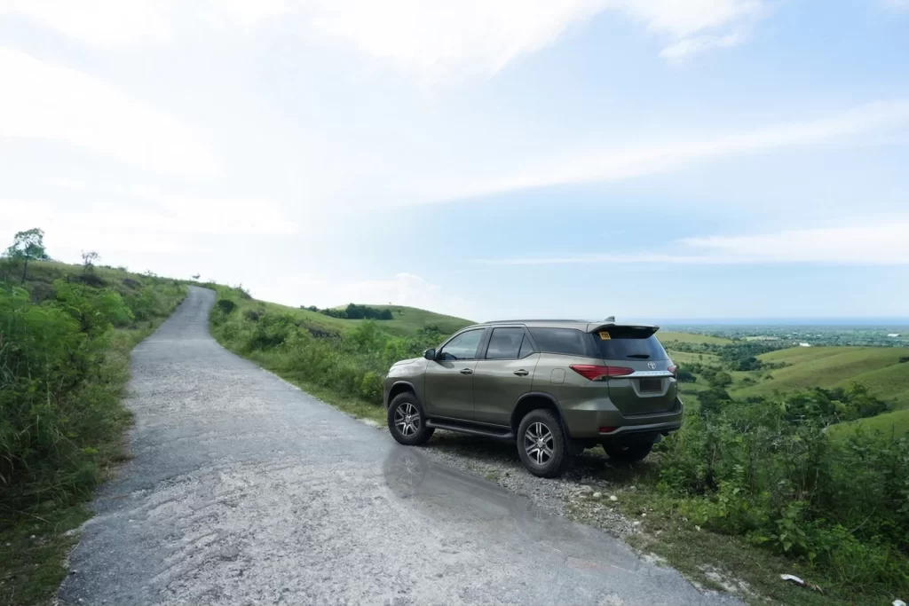 Olive green SUV is parked on the side of a paved road, overlooking green hills under a blue and white sky.