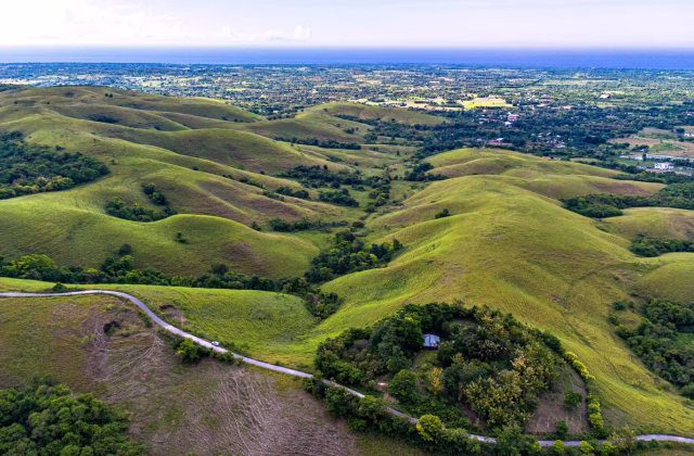 Aerial view shows green rolling hills and valleys on Sumba Island, with a road and a few buildings visible. Sumba geography.