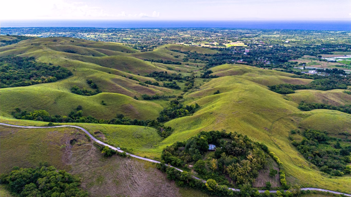 Aerial view shows green rolling hills and valleys on Sumba Island, with a road and a few buildings visible. Sumba geography.