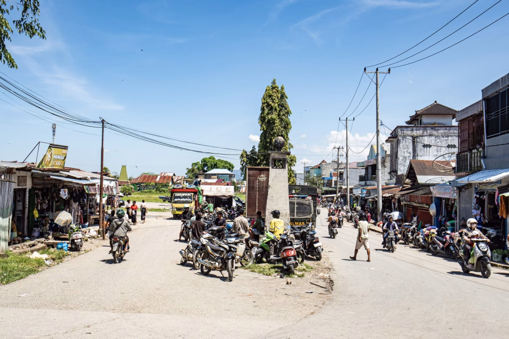 A street scene in Sumba, Indonesia shows motorbikes, small shops, and people under a bright blue sky.